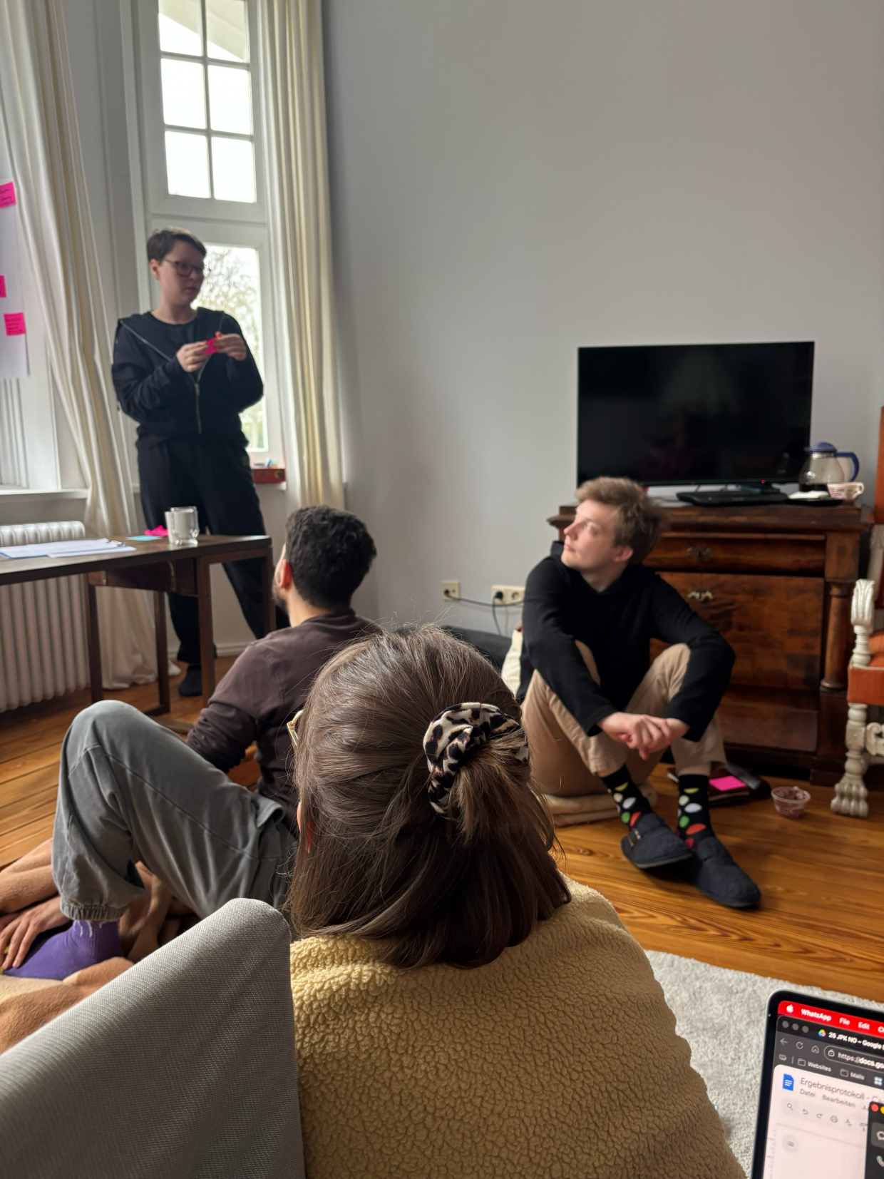 Gruppe von acht Personen bei einem Workshop im Wohnzimmer, sitzend im Kreis auf dem Boden; zwei Personen stehen am Fenster und ordnen bunte Klebezettel auf zwei Plakaten mit den Überschriften "Rahmenbedingungen" und "Themen"