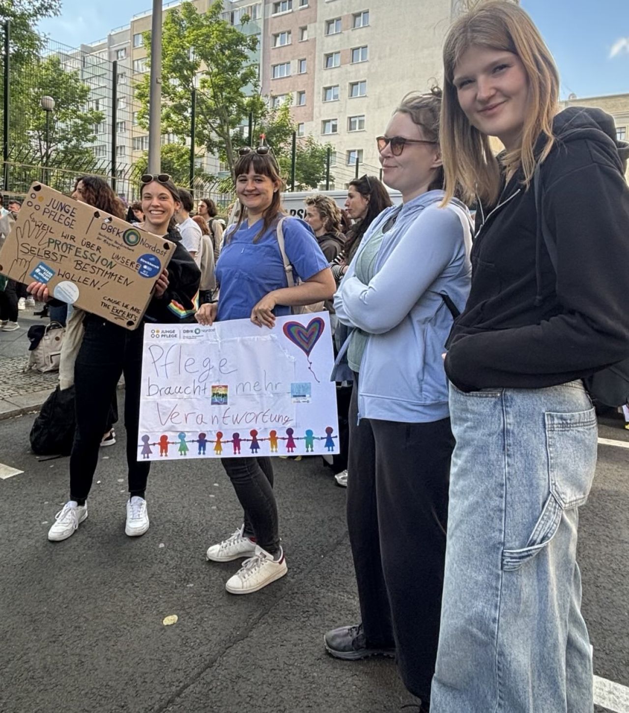 Das Bild zeigt eine Gruppe von jungen Frauen, die an einer Demonstration auf einer Straße teilnehmen. Sie stehen nebeneinander und schauen teilweise lächelnd in die Kamera. Im Hintergrund sind viele weitere Menschen zu sehen, die ebenfalls an der Veranstaltung teilnehmen. Außerdem erkennt man Bäume, eine Straßenlaterne und mehrstöckige Gebäude in einer städtischen Umgebung.  Zwei der Frauen halten selbstgemachte Plakate hoch. Auf einem steht sinngemäß, dass **junge Pflegekräfte mehr über ihre eigene Profession selbst bestimmen wollen**. Auf dem anderen Plakat steht **„Pflege braucht mehr Verantwortung“**, dazu sind bunte Figuren und ein Herz gezeichnet. Auf den Schildern sind auch Logos von Pflege-Organisationen zu sehen.  Die Teilnehmerinnen wirken wie Studierende oder junge Pflegekräfte. Eine Person trägt beispielsweise eine blaue medizinische Arbeitskleidung, was darauf hindeutet, dass sie im Gesundheitsbereich arbeitet. Die Stimmung wirkt freundlich und solidarisch, gleichzeitig vermittelt die Szene Engagement für bessere Bedingungen oder mehr Mitbestimmung in der Pflege.  Insgesamt zeigt das Bild eine **friedliche Demonstration von jungen Menschen aus dem Pflegebereich**, die sich mit kreativen Plakaten für Veränderungen und mehr Einfluss ihrer Berufsgruppe einsetzen.